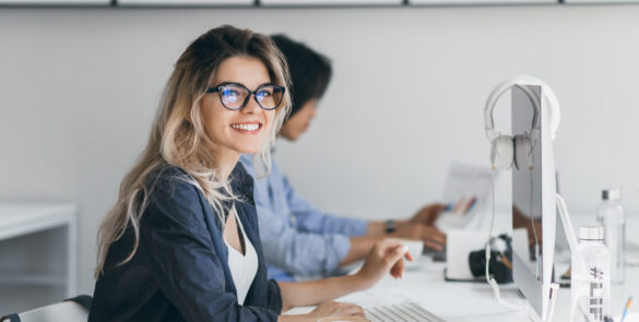 Attractive laughing freelancer girl posing with cup of coffee at her workplace. Chinese student in blue shirt works with document in campus with blonde friend in glasses.